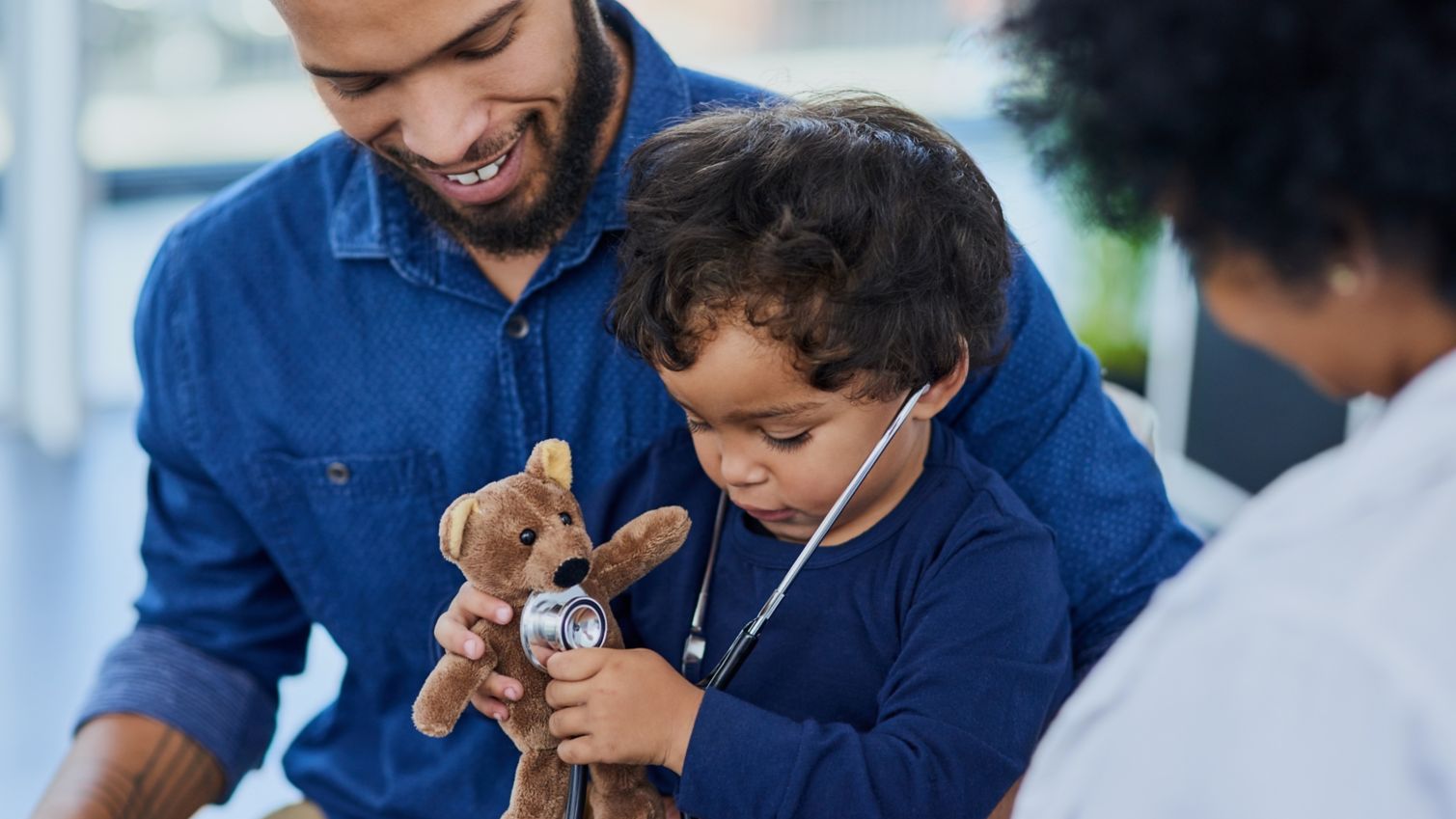 Young boy examines teddy bear while dad and doctor watch